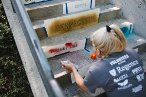 A woman volunteer spray paints the phrase "you got this" with a stencil on the side of a concrete step
